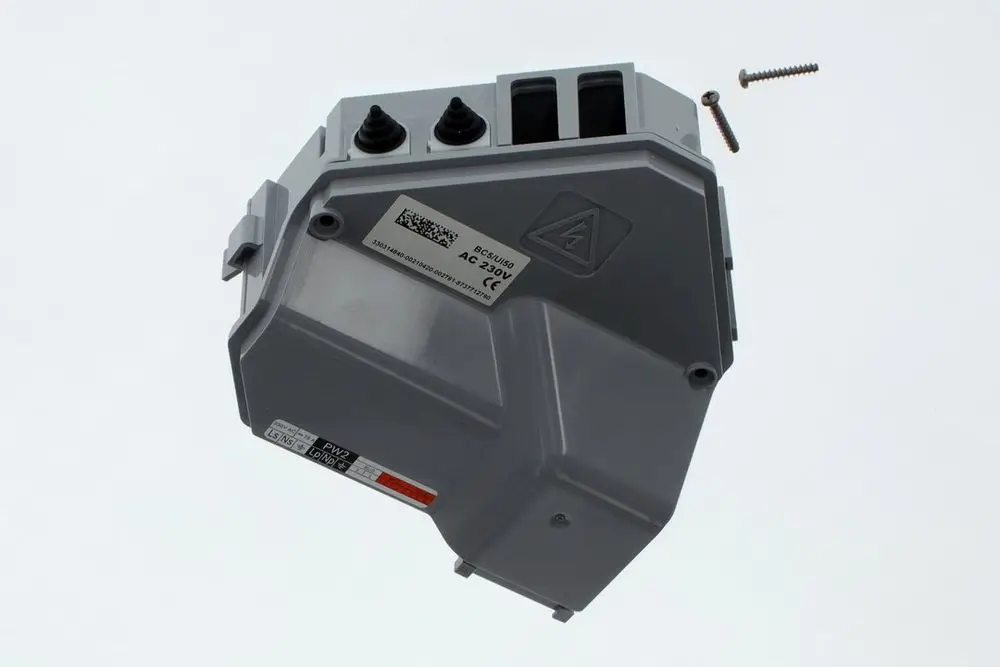 Graue Bosch I/O-Box, die an einer Unterseite sichtbare Anschlüsse und eine Warnung vor elektrischer Gefahr zeigt. Einzelne Schraube liegt daneben.